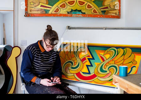 Woman artist working sitting by a drawing in progress at drawing table in a workshop, checking her mobile phone. Stock Photo