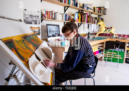 Woman artist sitting at a tilted drawing table in her studio, drawing, using several sheets of stiff paper. Stock Photo