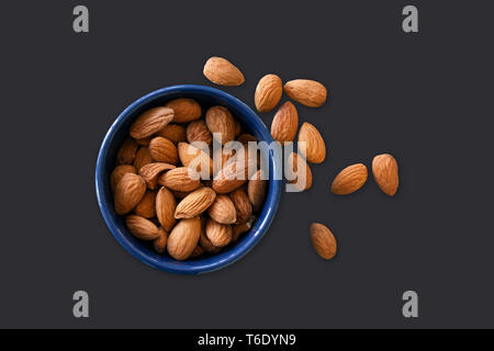 Almonds lying in a blue bowl isolated on dark background with almonds scattered around. Top view image Stock Photo