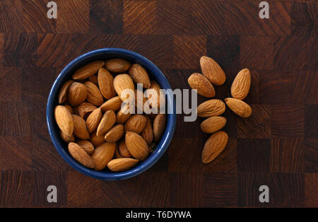 Almonds lying in a blue bowl on dark wooden background with almonds scattered around. Top view image Stock Photo