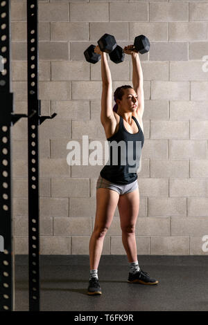 Fit strong young woman doing weightlifting exercises raising a pair of dumbbells above her head inside a gymnasium Stock Photo