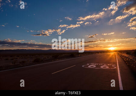 Route 66 pavement sign in the foreground and the diminishing perspective of the road leading to a dramatic sunset in the Mojave desert outside of Ambo
