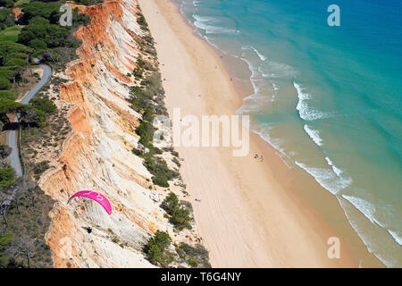 AERIAL VIEW. Paraglider using the sea breeze to soar alongside a colorful seaside cliff. Praia da Falésia, Albufeira, Algarve, Portugal. Stock Photo