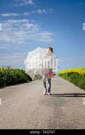 little girl with umbrella walking among yellow rapeseed fields in empty rural path Stock Photo