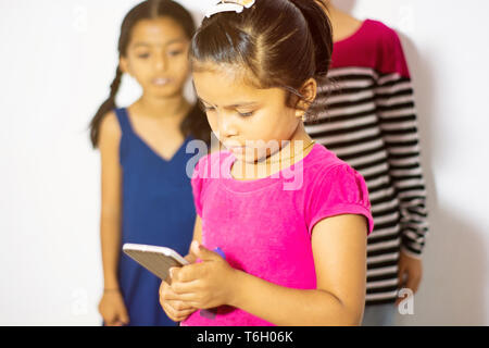 Cute Indian little kid using mobile device and two kids watching the phone from her behind Stock Photo