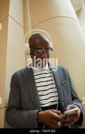 Waist up portrait of trendy African man wearing headphones using smartphone standing by architectural building, copy space Stock Photo