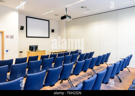 Empty presentation room with rows of blue seats, white projection screen, video projector on the ceiling, computer screen on a desk and white walls. Stock Photo