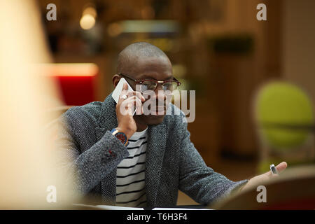 Portrait of contemporary African-American businessman sitting at table and speaking by smartphone, copy space Stock Photo