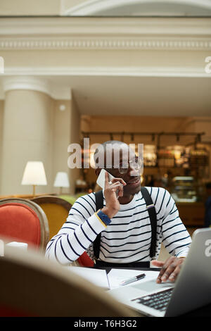 Portrait of contemporary African man speaking by phone and  using laptop while studying in library, copy space Stock Photo