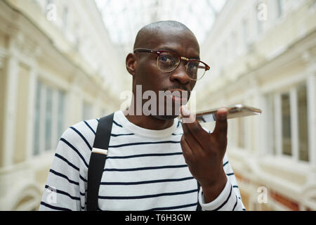 Portrait of contemporary African man speaking by phone on speaker standing in large architectural building, copy space Stock Photo