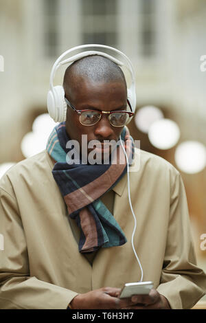 Portrait of trendy African man wearing headphones and using smartphone while posing in beautiful building outdoors, copy space Stock Photo