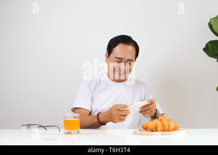 Happy senior couple having croissant breakfast at home and drinking orange juice, using smartphone. Stock Photo