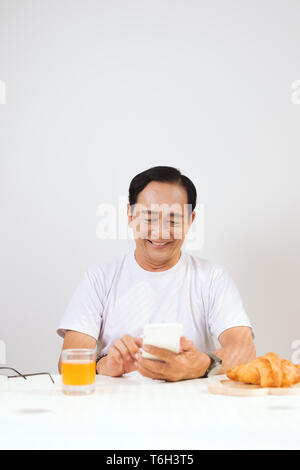 Happy senior couple having croissant breakfast at home and drinking orange juice, using smartphone. Stock Photo