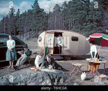 1950s camping. A family is enjoying their holiday and the practial camping life in their caravan. Demonstrating how well everything works for them even on holiday. The mother holds out a pot with a ready cooked meal while her daughter has set the table. The father and daughter spends their time looking at a map probably to find the next suitable location and campsite. Everything in this pictures illustrates very well the time of the 1950s both in visible objects and clothing. Other aspects of the time is that while the women is pictured cooking, the man is not. Note the model of the caravan an Stock Photo