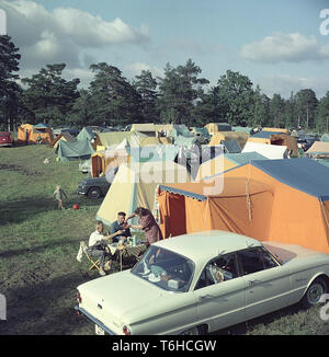 Camping in the 1950s. A camping ground where people spend parts of their summer holiday. The grounds are full of tents in different sizes. Beside the tents their cars are parked. In front a group of three people having a cup of coffee sitting in the typical folding camping chairs and table. Sweden 1958 Stock Photo