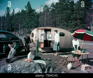 1950s camping. A family is enjoying their holiday and the practial camping life in their caravan. Demonstrating how well everything works for them even on holiday. The mother holds out a pot to use for the  meal while her daughter is setting the table.  Everything in this pictures illustrates very well the time of the 1950s both in visible objects and clothing. Other aspects of the time is that while the women is pictured cooking, the man is not. Note the model of the caravan and the special feature with a lowered floor that makes the caravan more spacious. This never returned in future carava Stock Photo