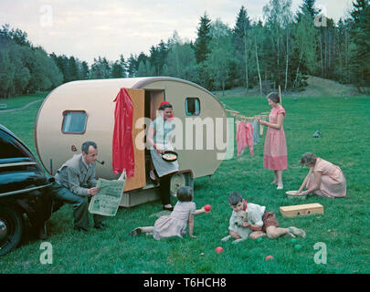 1950s camping. A family is enjoying their holiday and the practial camping life in their caravan. Demonstrating how well everything works for them even on holiday. The mother has fried some eggs and and steps out of the caravan holding the pan. Her eldest daughters are washing dishes and hanging up the laundry to dry. The youngest children are playing and the father of course sits around doing nothing more then having a smoke and reading the newspaper as a typical 1950s father would do.Everything in this pictures illustrates very well the time of the 1950s both in visible objects and clothing. Stock Photo