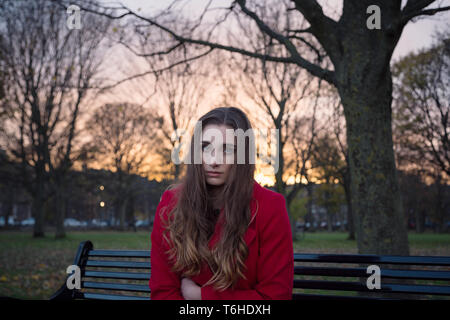 A young woman sitting on a bench in a park, with lots on her mind, feeling isolated and anxious. Stock Photo