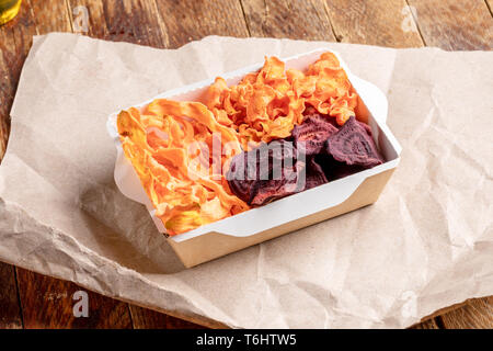 Paper box with vegetable chips on wrapping paper on old wooden table. Stock Photo