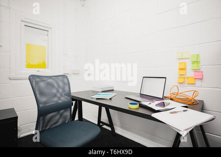 A shot of a wooden desk in an empty office, on the desk is a computer, paperwork and a smart phone. Post-it notes can be seen on the wall of the offic Stock Photo