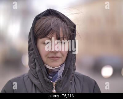 Young brunette woman in the hood on the background of a city street, stands with eyes closed Stock Photo