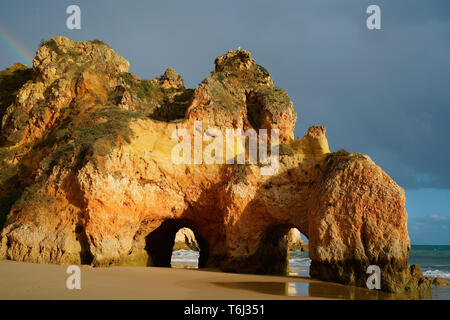 Double sea arch under dark and stormy clouds, small rainbow in the upper left. Near Prainha Beach, Alvor, Algarve, Portugal. Stock Photo