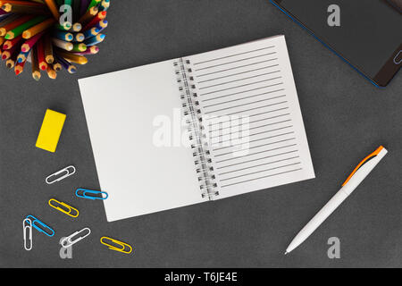 Open white line spiral paper notebook with mobile phone, pen, colored pencils, eraser and paper clips on empty dark background top view Stock Photo