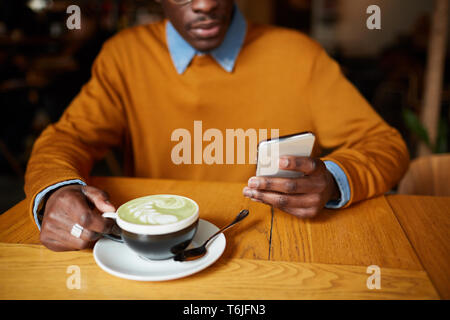 Warm toned shot of contemporary African-American man using smartphone while sitting at table in coffee shop, copy space Stock Photo