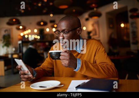 Warm toned portrait of contemporary African-American man using smartphone sitting at table in coffee shop, copy space Stock Photo