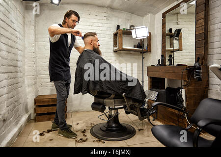 Brutal guy in modern Barber Shop. Hairdresser makes hairstyle a man with a long beard. Master hairdresser does hairstyle by scissors and comb Stock Photo