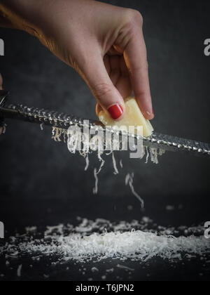 Hand of a woman grating parmesan cheese on a black background. Dark food. Italian cheese Parmigiano Reggiano Stock Photo