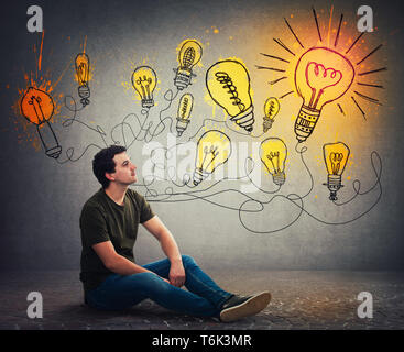 Relaxed man sitting on the floor smiling looking at bright lightbulbs on the wall. Smart and ingenious guy has different thinking, genius creativity c Stock Photo