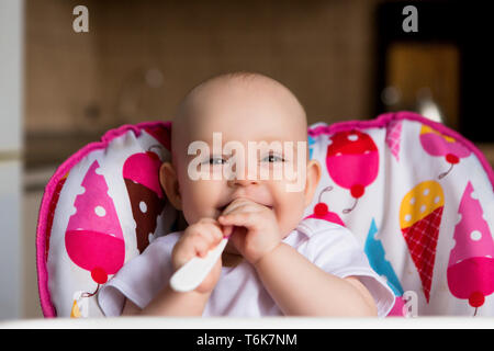 Baby in the kitchen in a baby chair holding a spoon and smiling.Sweet baby in high chair.Cheerful baby child eats food itself with spoon. Stock Photo