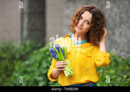 Happy young fashion woman with bouquet of flowers walking in city street Stylish female model wearing yellow shirt and blue skirt Stock Photo