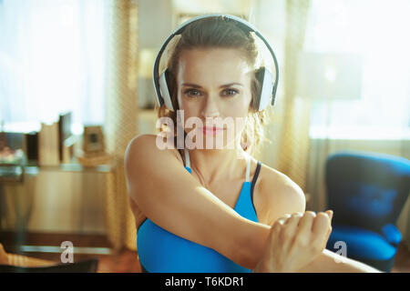 Portrait of healthy sports woman in fitness clothes listening to the music with headphones and stretching at modern home. Stock Photo