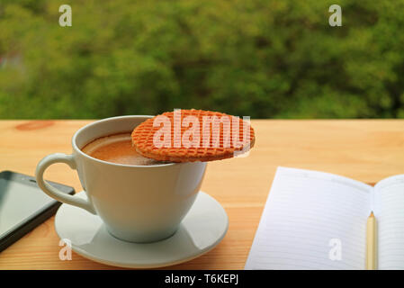 Cup of Hot Coffee with a Stroopwafel Placed on Top with Lined Note Papers and Cellphone on Wooden Table Stock Photo