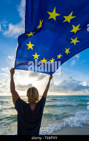 Man with blond hair holding EU European Union flag flying in the wind on a sunrise Mediterranean Beach Stock Photo