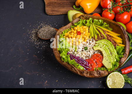 Wooden bowl with chickpea, avocado, wild rice, quinoa, bell pepper, tomatoes, greens, cabbage, lettuce on dark table and wooden spoon with chia seeds  Stock Photo