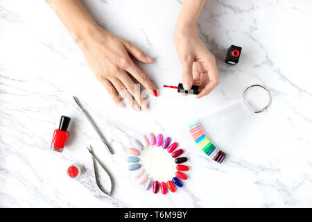 Beautiful woman hands painting nails with red nail polish on marble table with manicure set on it, top view. Stock Photo