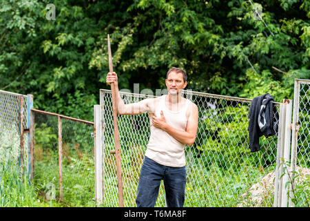 Man in garden cutting weeds oat grass with sickle scythe manual tool in green summer in Ukraine dacha or farm farmer hot scratching armpit Stock Photo