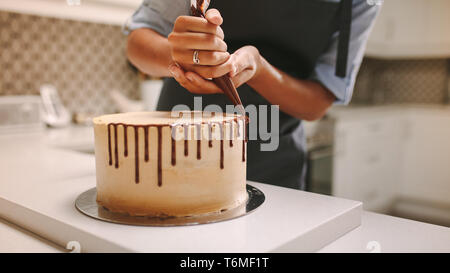 Close up of hands of a female chef with confectionery bag squeezing liquid chocolate on cake. Pastry chef decorating a cake in kitchen. Stock Photo
