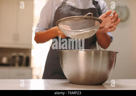 Close up of female pastry chef sifting flour in a bowl to make dough in kitchen. Woman baker baking in home kitchen. Stock Photo