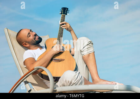 Portrait of a spanish man with beard and short buzz hair, playing an acoustic guitar outdoors over a blue sky background. Stock Photo