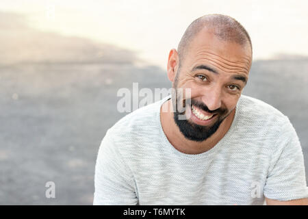 Portrait of a spanish man with a beard and ultra-short buzz, smiling in a funny way, looking camera, wearing a t-shirt, outdoors. Stock Photo