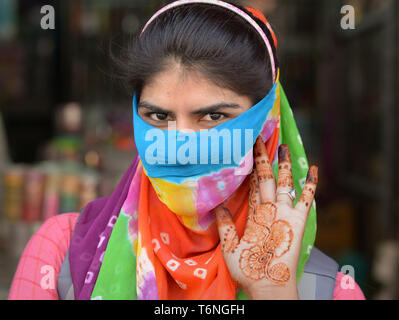 Young Indian Rajasthani woman covers her face with a colourful secular dust veil and shows the intricate henna tattoos on her left palm. Stock Photo