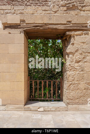 Rectangular opening with wooden balustrade on old abandoned stone bricks wall revealing part of a big green dense tree in summer time Stock Photo