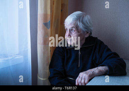 An old woman looks longingly out the window of her house. Care for lonely elderly. Stock Photo