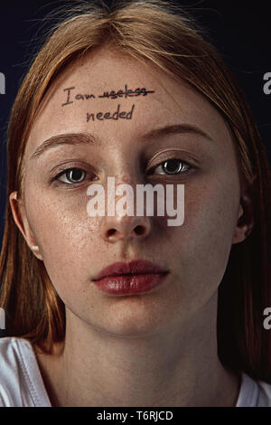 Portrait of young woman with mental health problems. The image of a tattoo on the forehead with the words I am useless-needed. Concept of hidding the true feelings, psycological trouble, treatment. Stock Photo