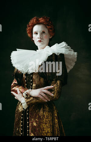 Too serious. Medieval redhead young woman in golden vintage clothing as a duchess standing crossing hands on dark green background. Concept of comparison of eras, modernity and renaissance. Stock Photo