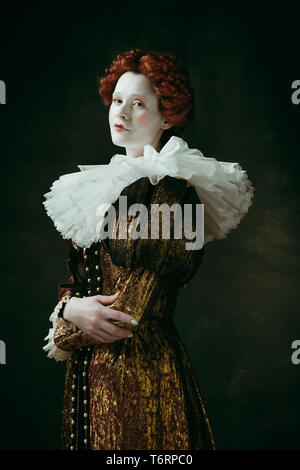 Lovely portrait. Medieval redhead young woman in golden vintage clothing as a duchess standing crossing hands on dark green background. Concept of comparison of eras, modernity and renaissance. Stock Photo